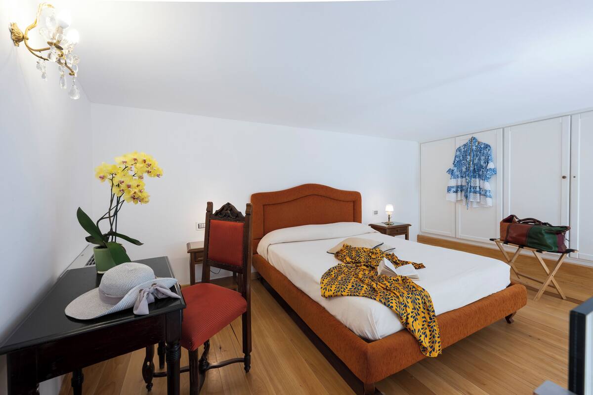A hotel room with a wooden floor, a single bed with a white and orange headboard, a desk with a chair, a small table with a plant, and a wardrobe with clothes hanging inside.