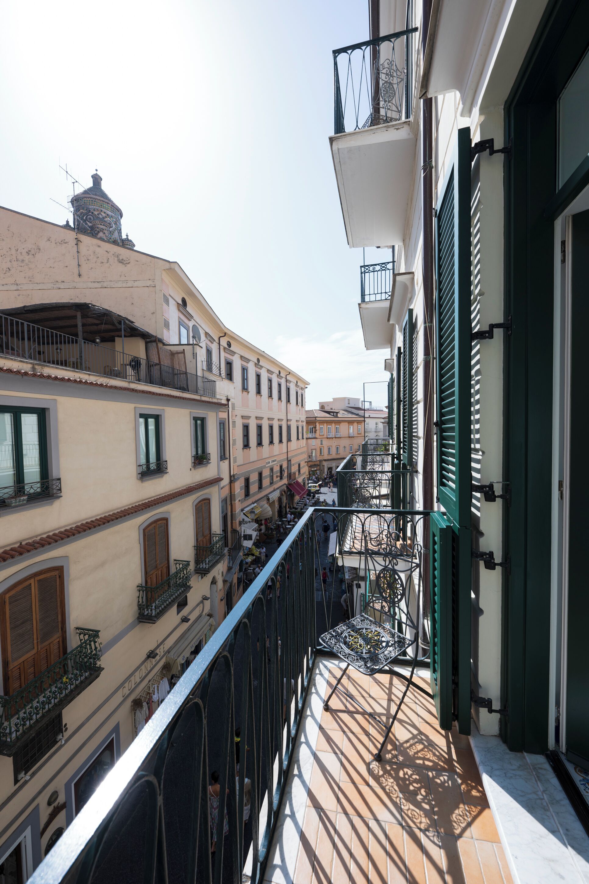 Una estrecha calle urbana flanqueada por edificios, con balcones de barandilla metálica y cielo despejado.