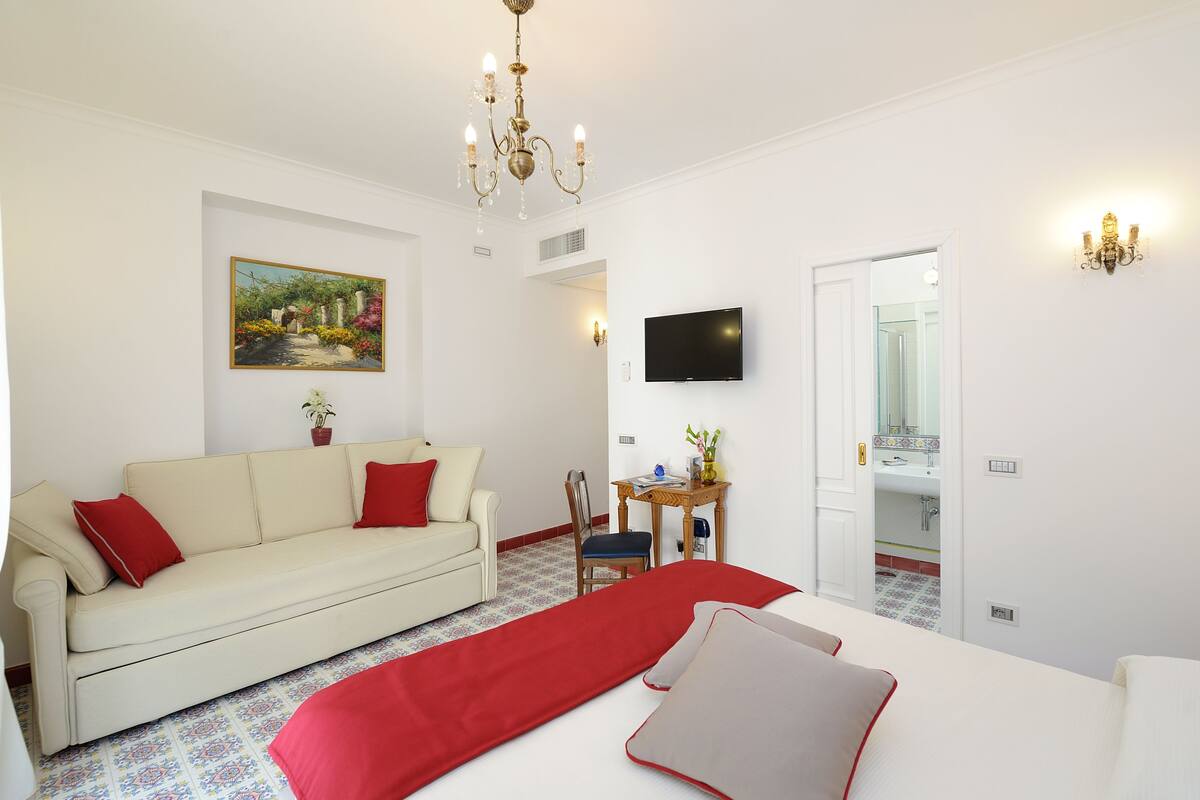 A hotel room with a white sofa, red cushions, a red bedspread, a TV mounted on the wall, a wooden side table, and a painting of a garden.