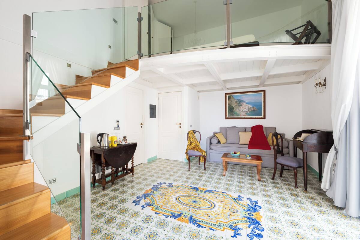 A spacious living room with a wooden staircase, a glass railing, a patterned rug, and a painting on the wall.