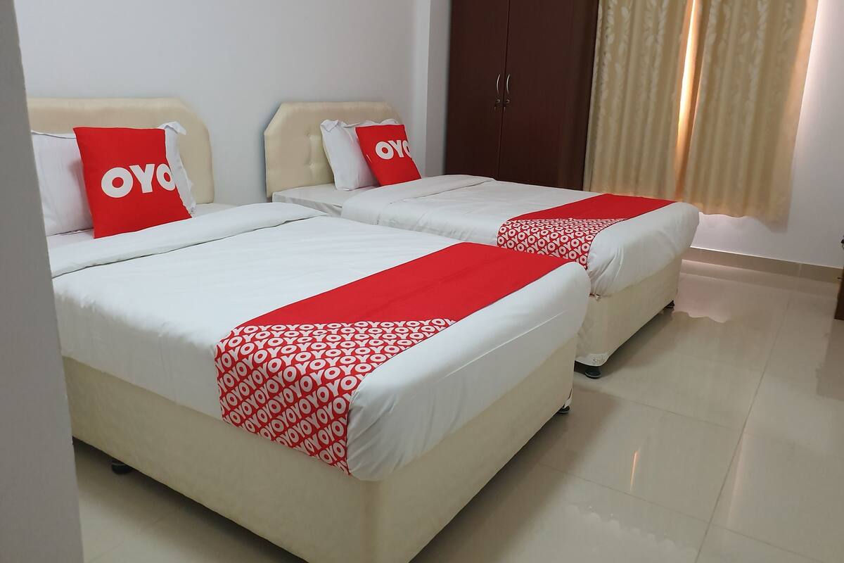 Two beds with white bedding and red accents, a wooden wardrobe, and a window with curtains.