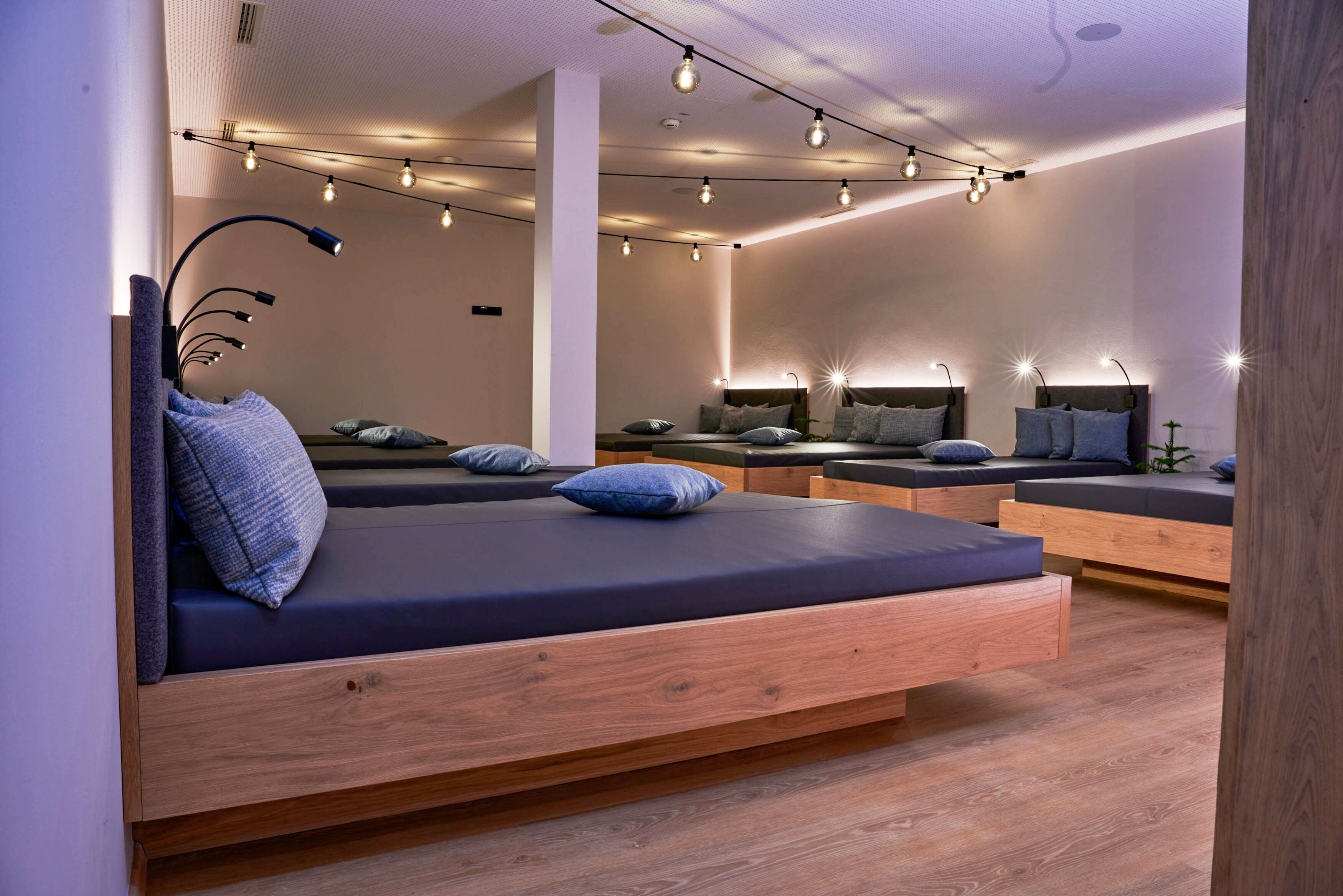 Une chambre moderne avec plusieurs lits, chacun doté d’un cadre en bois et d’une literie aux couleurs sombres. Des guirlandes lumineuses sont accrochées au plafond et un grand miroir orne le mur.