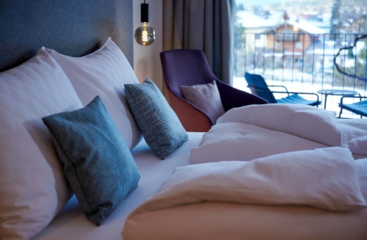 Una cama bien hecha con varias almohadas, una silla moderna y vistas al balcón.