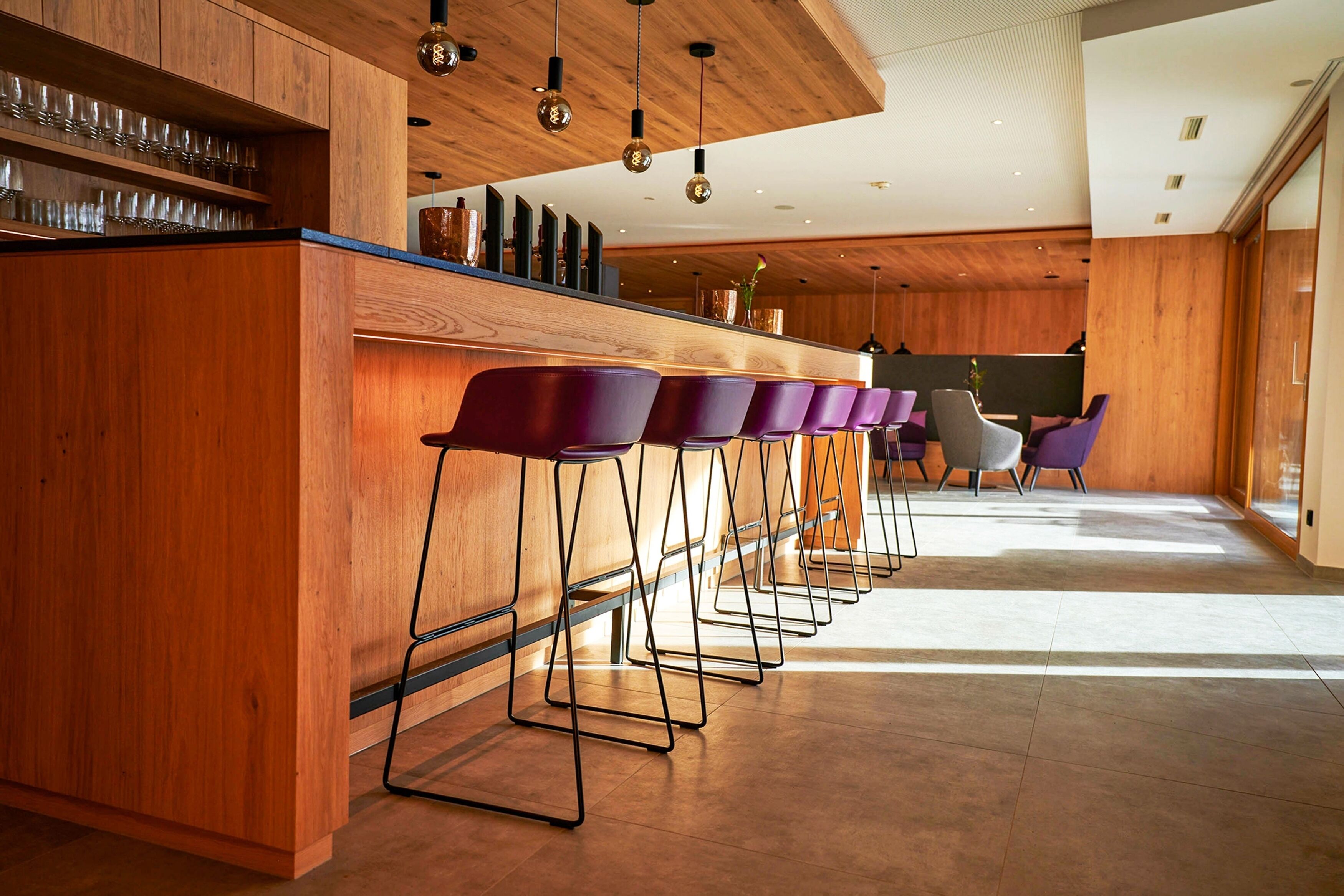 Un área de bar moderna con paneles de madera, una barra larga y varios taburetes de bar morados.
