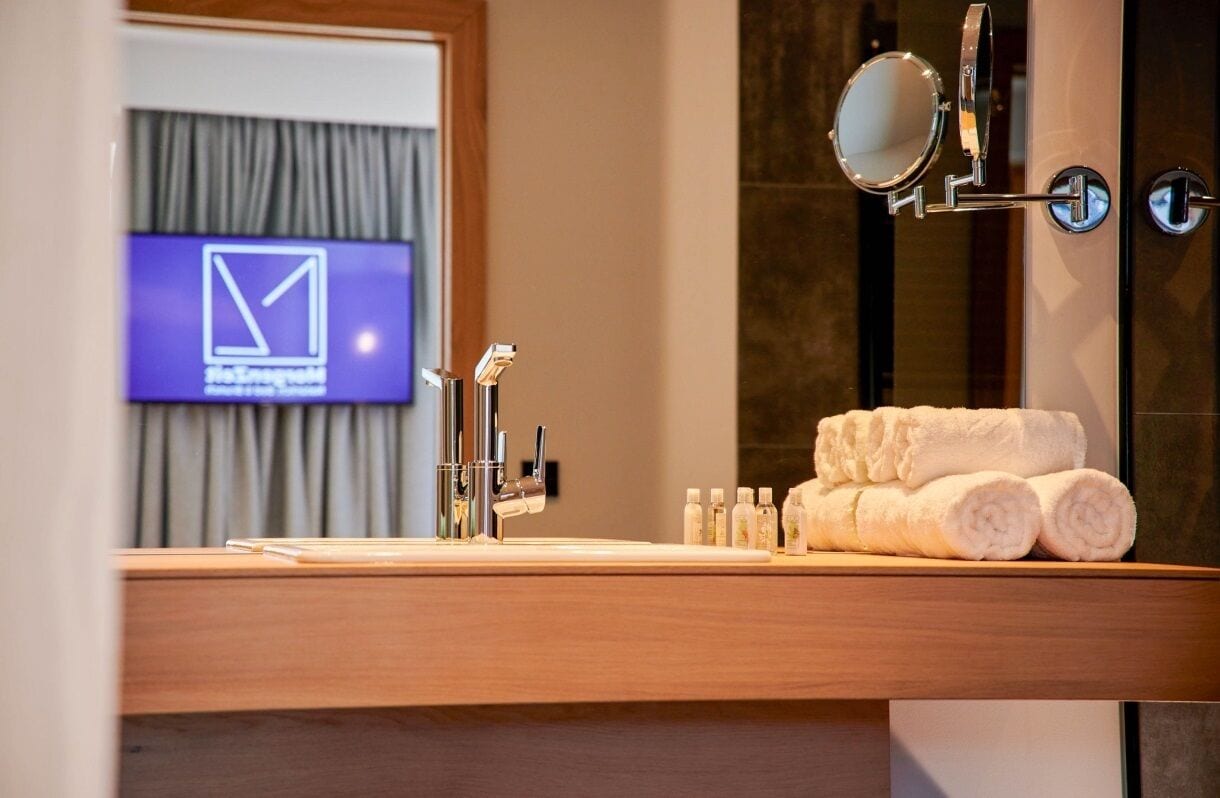 A hotel bathroom with a wooden vanity, a mirror, and a television displaying a logo.