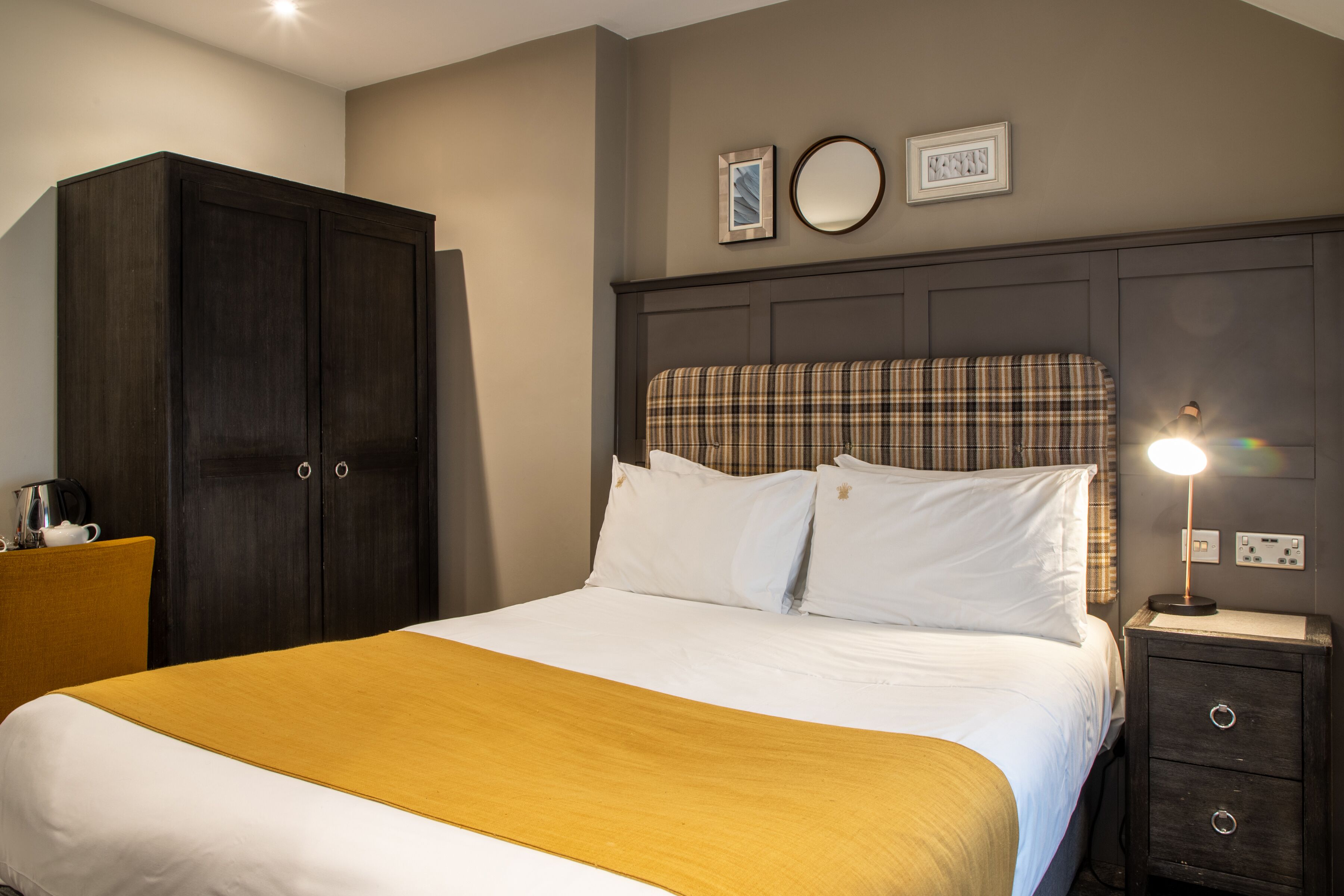 A neatly made bed with a plaid headboard, a bedside table with a lamp, and a wardrobe.