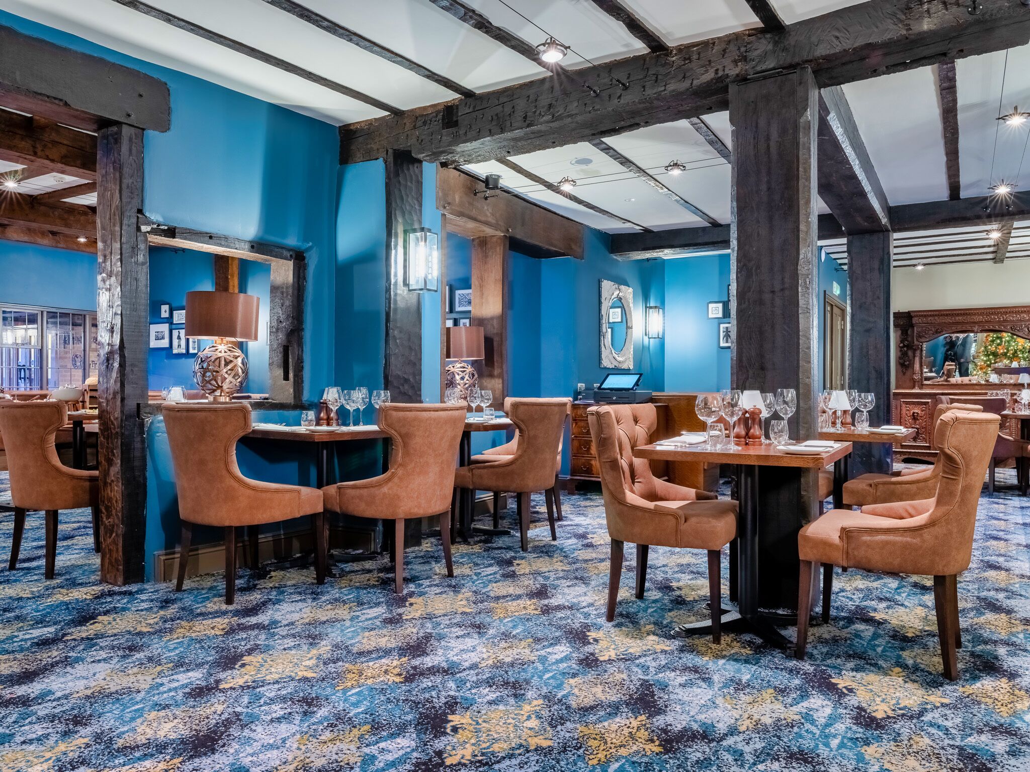 A restaurant with blue walls, wooden beams, and a patterned carpet. Tables are set with chairs and wine glasses.