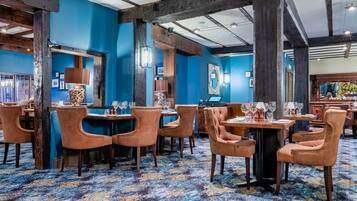 A restaurant with blue walls, wooden beams, and a patterned carpet. Tables are set with chairs and wine glasses.