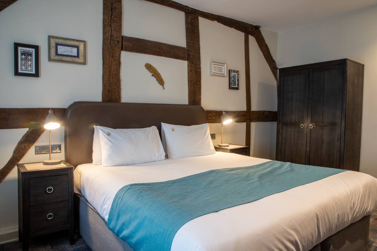 A bedroom with a large bed, wooden beams on the ceiling, two bedside tables with lamps, and a wardrobe.