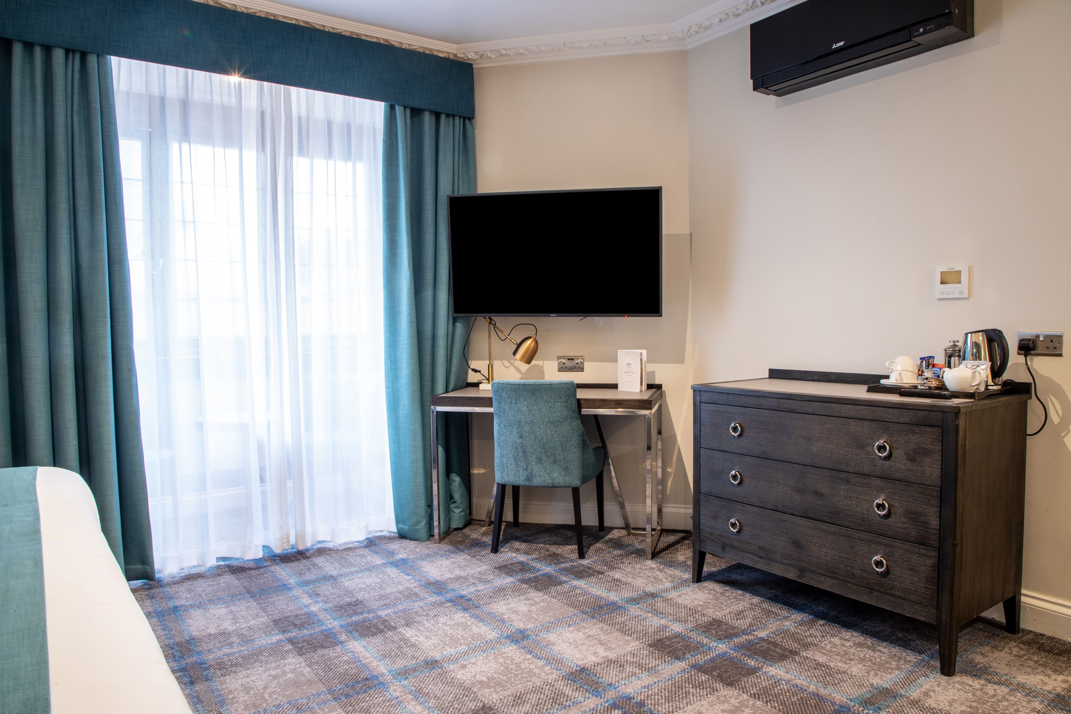 A hotel room with a bed, a desk with a chair, a flat-screen TV, a dresser, and a large window with curtains.