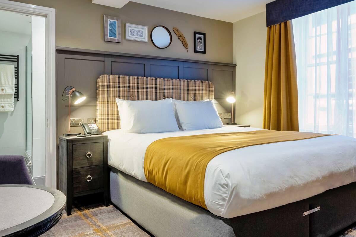 A neatly made bed with a plaid headboard, a bedside lamp, and a small table with a chair.