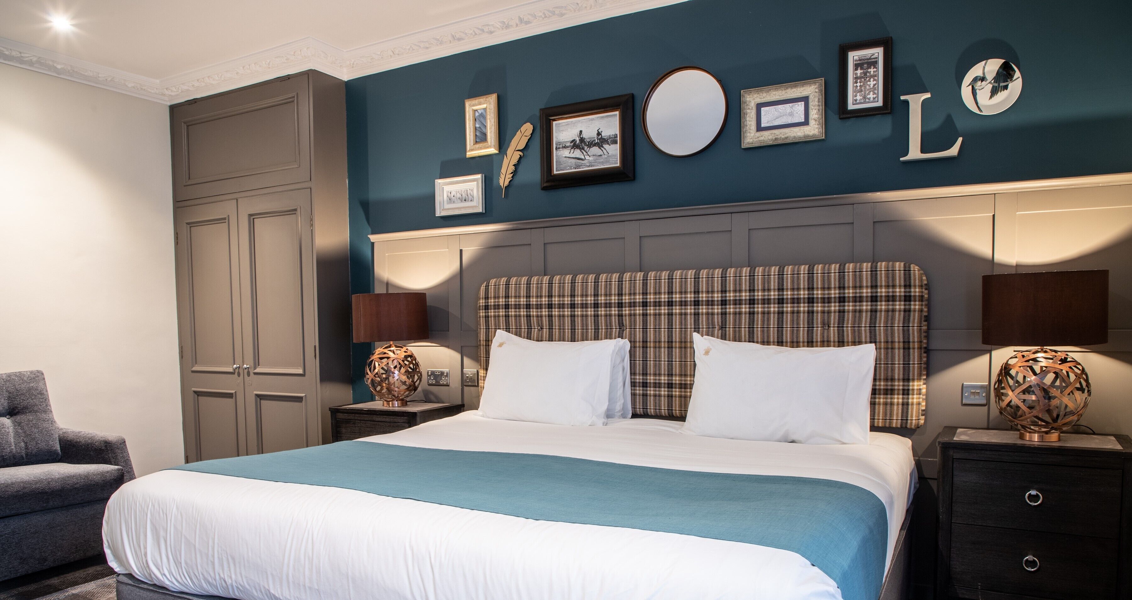 A neatly arranged hotel room with a large bed, a grey armchair, and a wall adorned with framed pictures and a mirror.