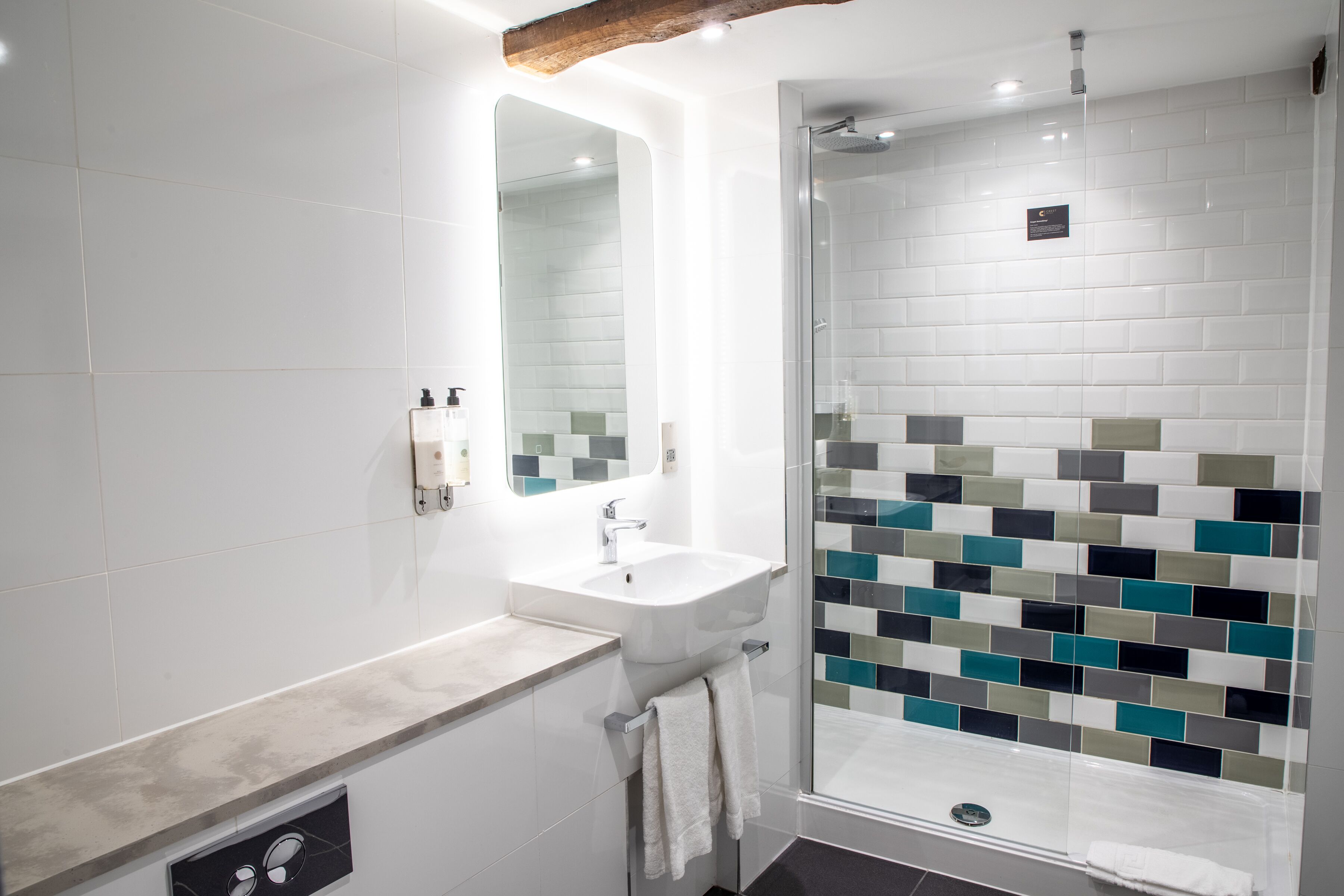A modern bathroom with a white sink, a mirror, and a glass-enclosed shower with multicolored tiles.