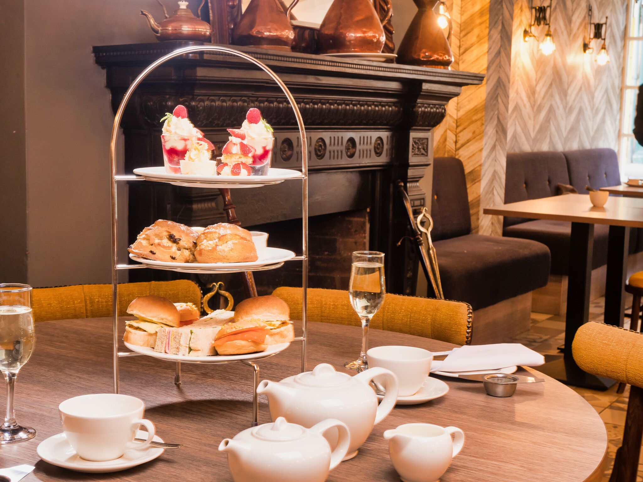 A three-tiered tea service stand with pastries and sandwiches, white teapots, cups, and a glass of water on a wooden table in a dining area with a fireplace and modern decor.