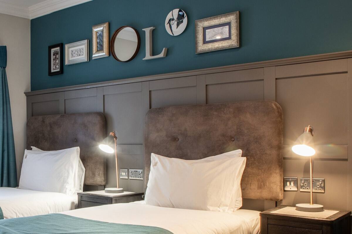 A hotel room with two beds, a large window with curtains, a headboard with a mirror, and framed pictures on the wall.