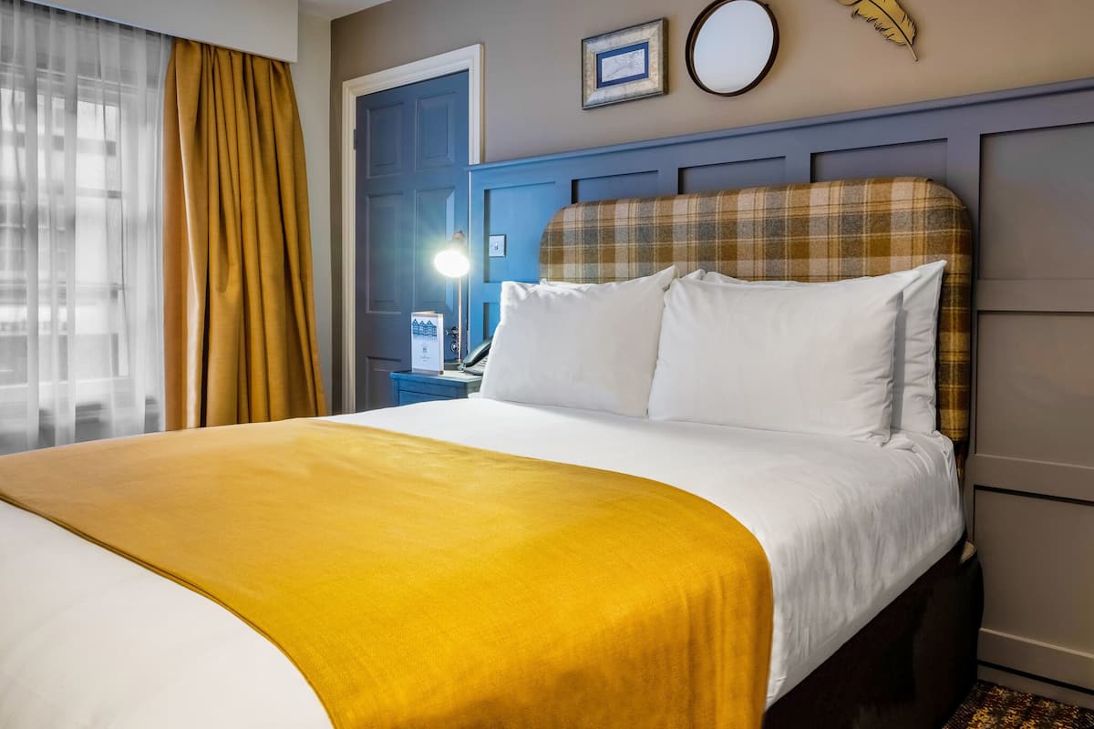 A hotel room with a bed, a nightstand, a lamp, a door, and a window with curtains.