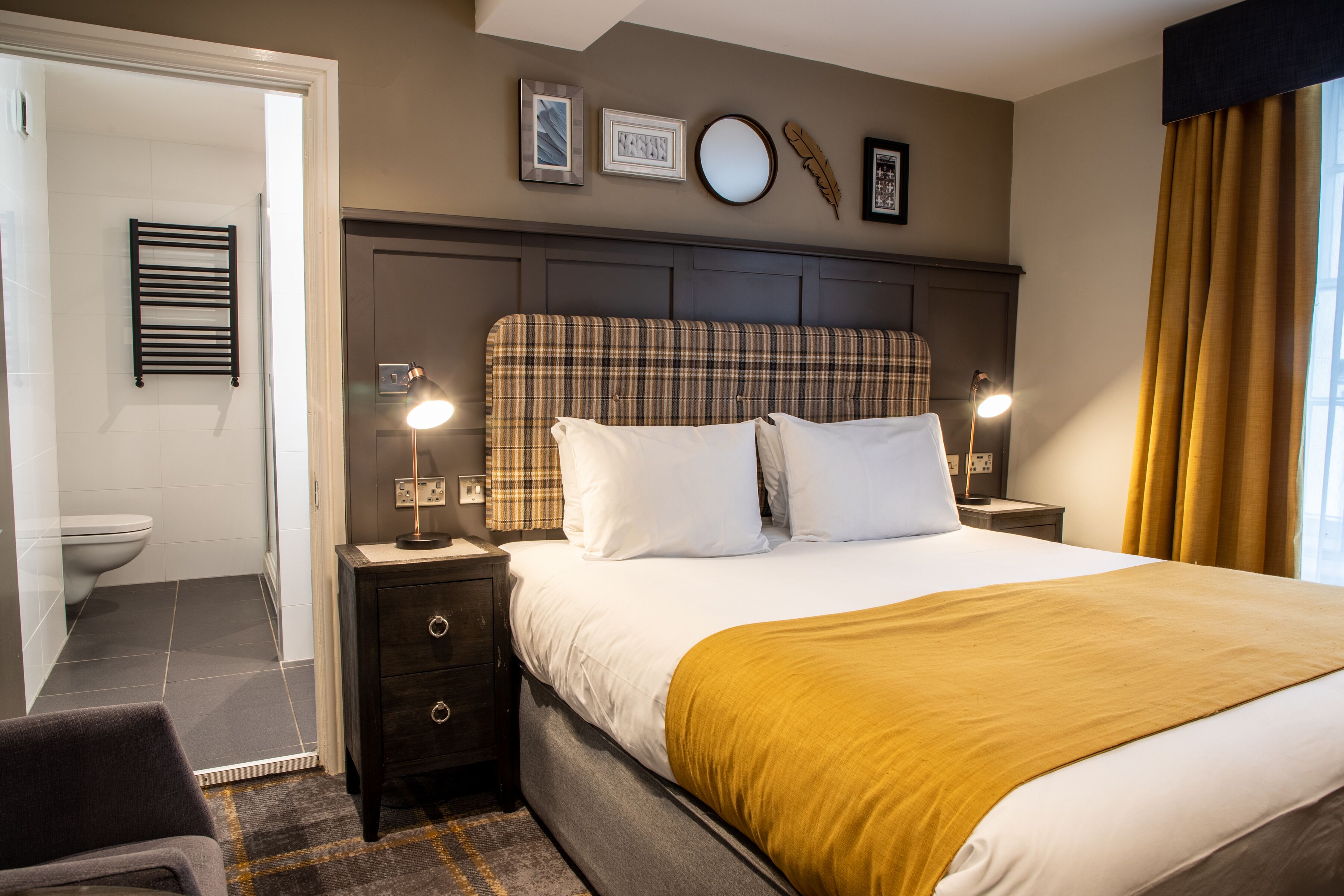 Hotelkamer met een bed, bedlampen, een hoofdbord, nachtkastje, een stoel en een zichtbare badkamer door een geopende deur.