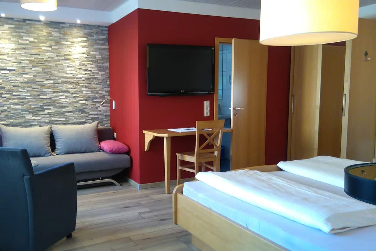 A hotel room with a stone wall, a red accent wall, a bed, a desk with a chair, and a television.