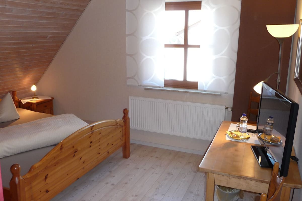 A small, cozy room with a sloped ceiling, a single bed, a wooden desk with a computer, and a window with patterned curtains.