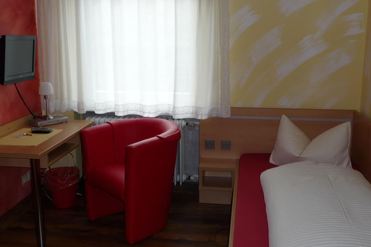 A hotel room with a bed, a red armchair, a desk with a television, and a window with sheer curtains.