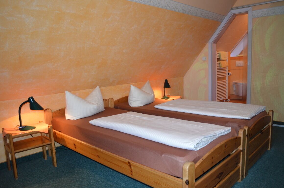 A bedroom with two wooden beds, each with white linens and pillows, a nightstand with a lamp, and a visible bathroom in the background.
