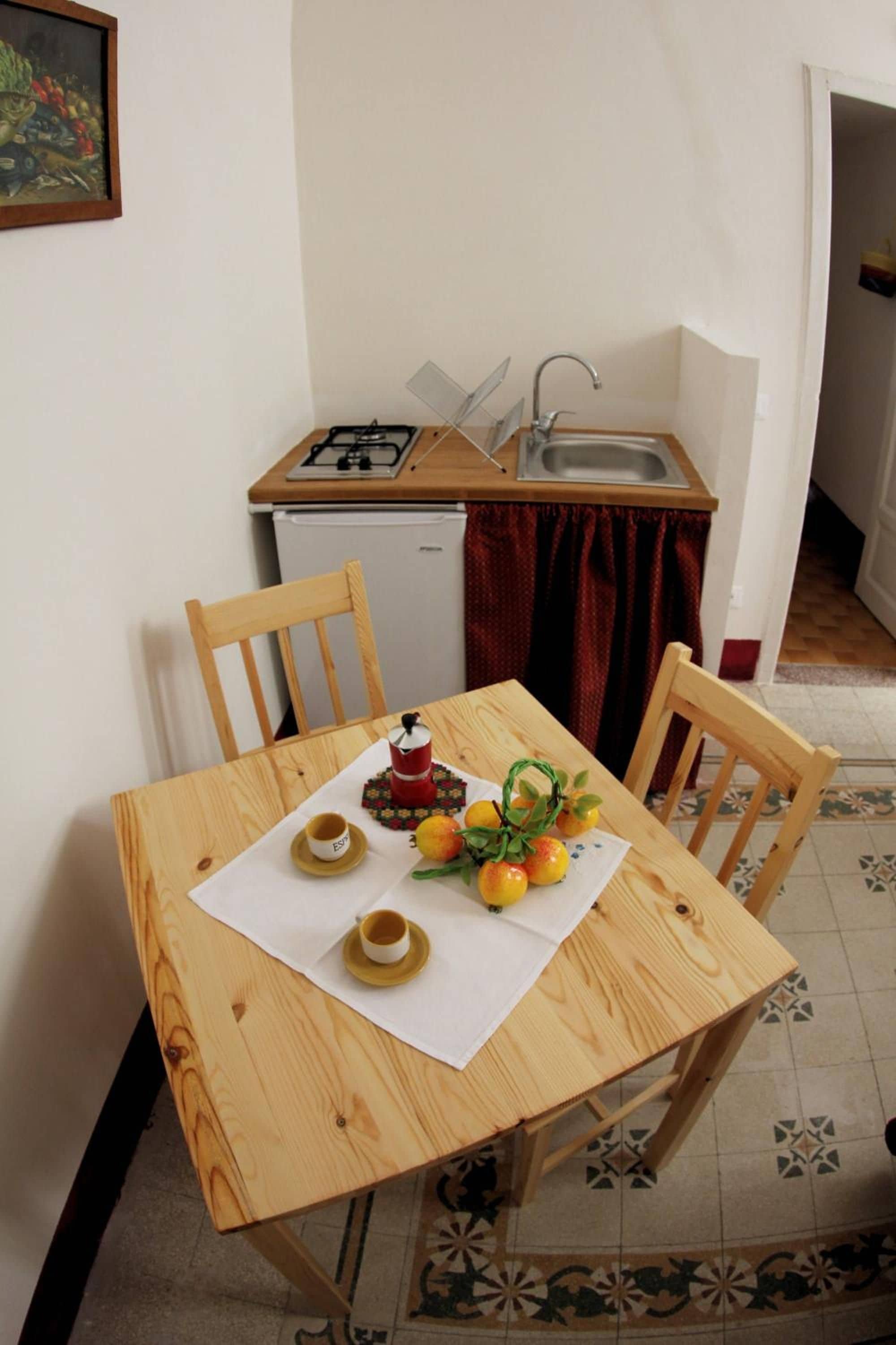 A small kitchen with a gas stove, a sink, and a refrigerator. There is a dining table with chairs and a small table setting with cups and lemons.