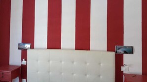 Una habitación con un cabecero blanco capitonado, paredes a rayas rojas y blancas, una mesita de noche pequeña con una lámpara y un cuadro en la pared.