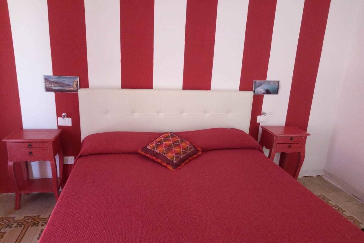 A bedroom with a red and white striped wall, a tufted headboard, a red bedspread, and two bedside tables with lamps.