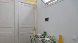 Una pequeña cocina con paredes blancas, una mesa de madera y una silla. Hay una ventana sobre la mesa y una puerta a la izquierda.