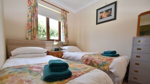 A hotel room with two beds, floral curtains, a nightstand with a lamp, a dresser with drawers, and a framed picture on the wall. - Stoats Old River Way (Winchelsea)