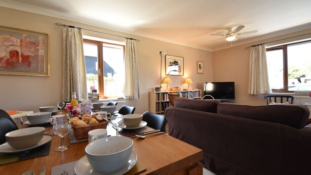 A dining area with a table set for breakfast, a sofa, a TV, and a ceiling fan. - Stoats Old River Way (Winchelsea)