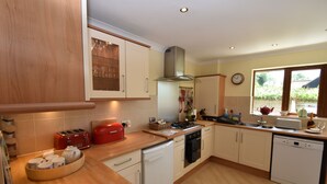 A modern kitchen with wooden cabinets, a red toaster, a dishwasher, and a window with a view of greenery. - Stoats Old River Way (Winchelsea)