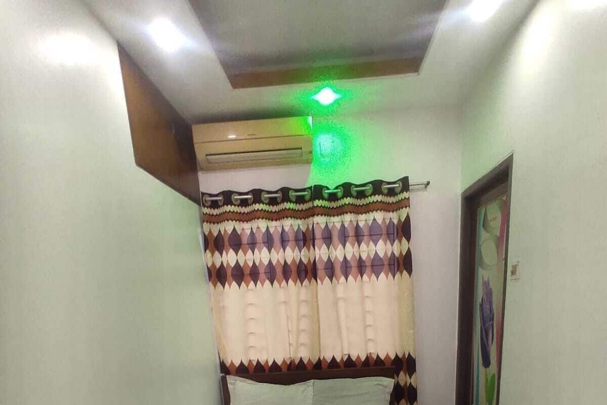 A small, neatly arranged bedroom with a single bed, a headboard with a patterned curtain, and a ceiling light.