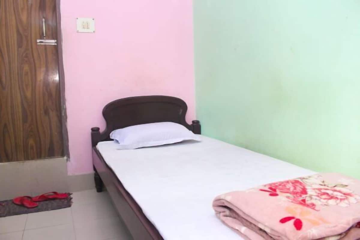 A single bed with a white pillow and a folded blanket, a wooden wardrobe, and a pair of red slippers on a tiled floor.