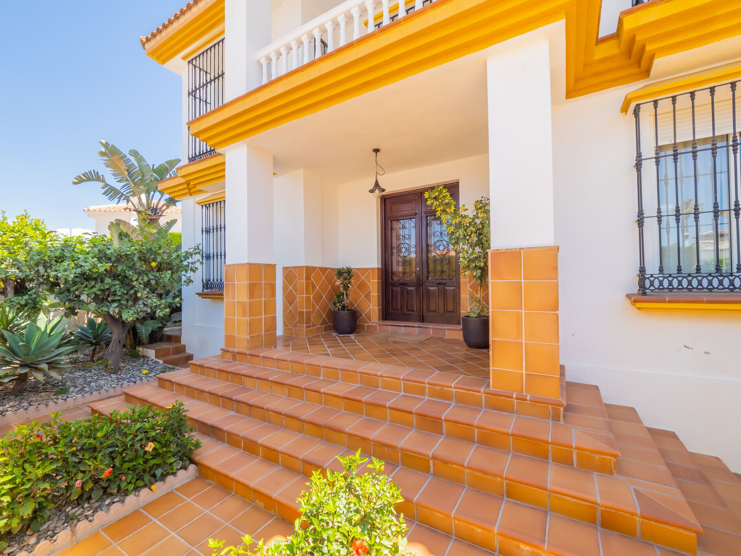 A two-story house with a tiled entrance, a balcony with yellow railings, and a door with a decorative iron grille.