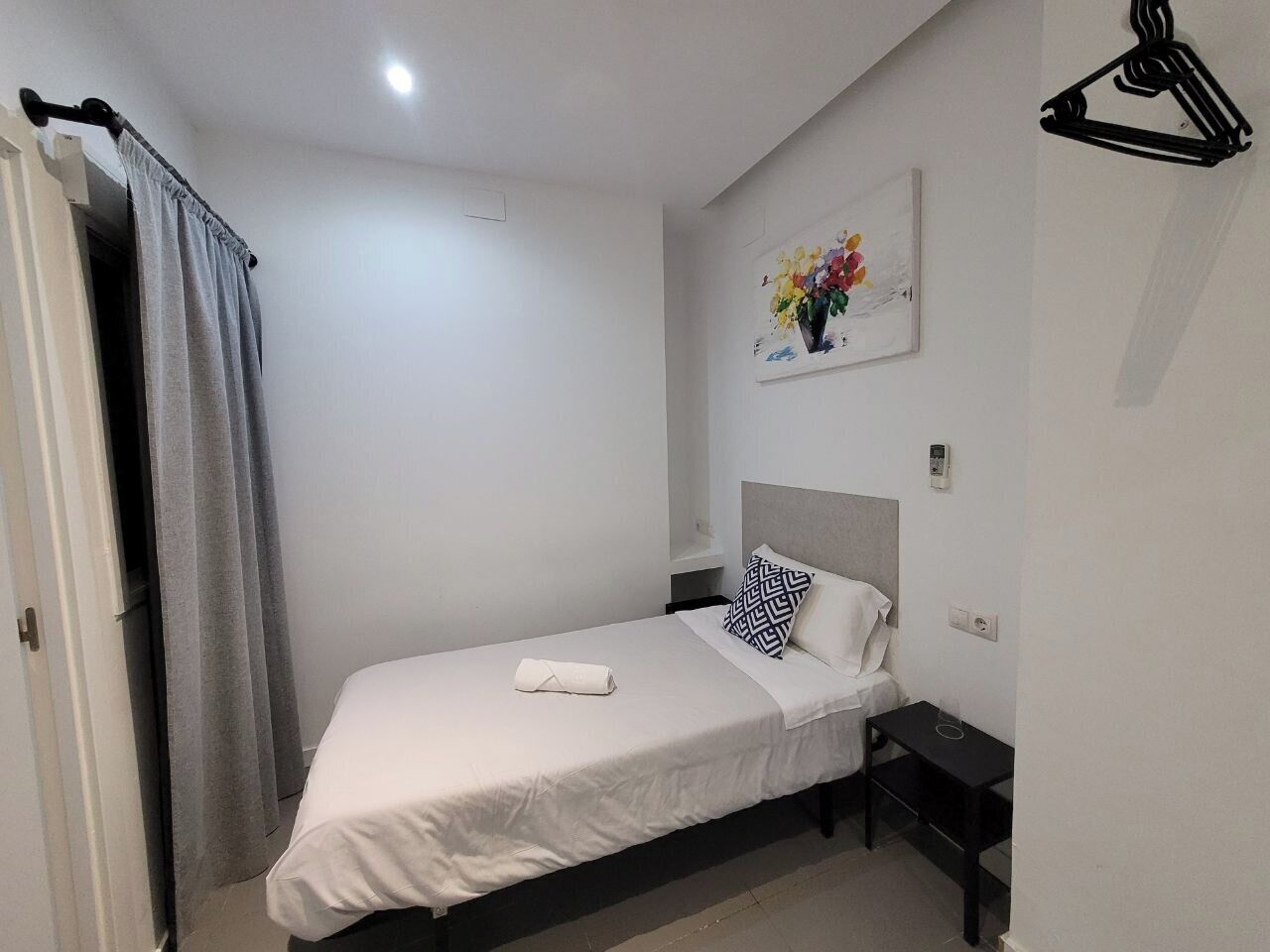 Una cama bien tendida con una colcha blanca y un cojín de estampado geométrico, una mesita de noche con una lámpara, y un cuadro de flores en la pared.