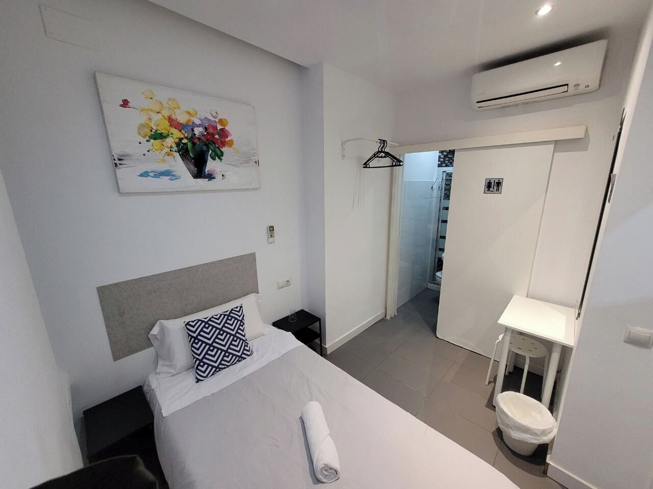 Een moderne hotelkamer met een net opgemaakt bed, een klein nachtkastje, een wandairconditioner, een deur met een bordje en een gang naar een andere kamer.