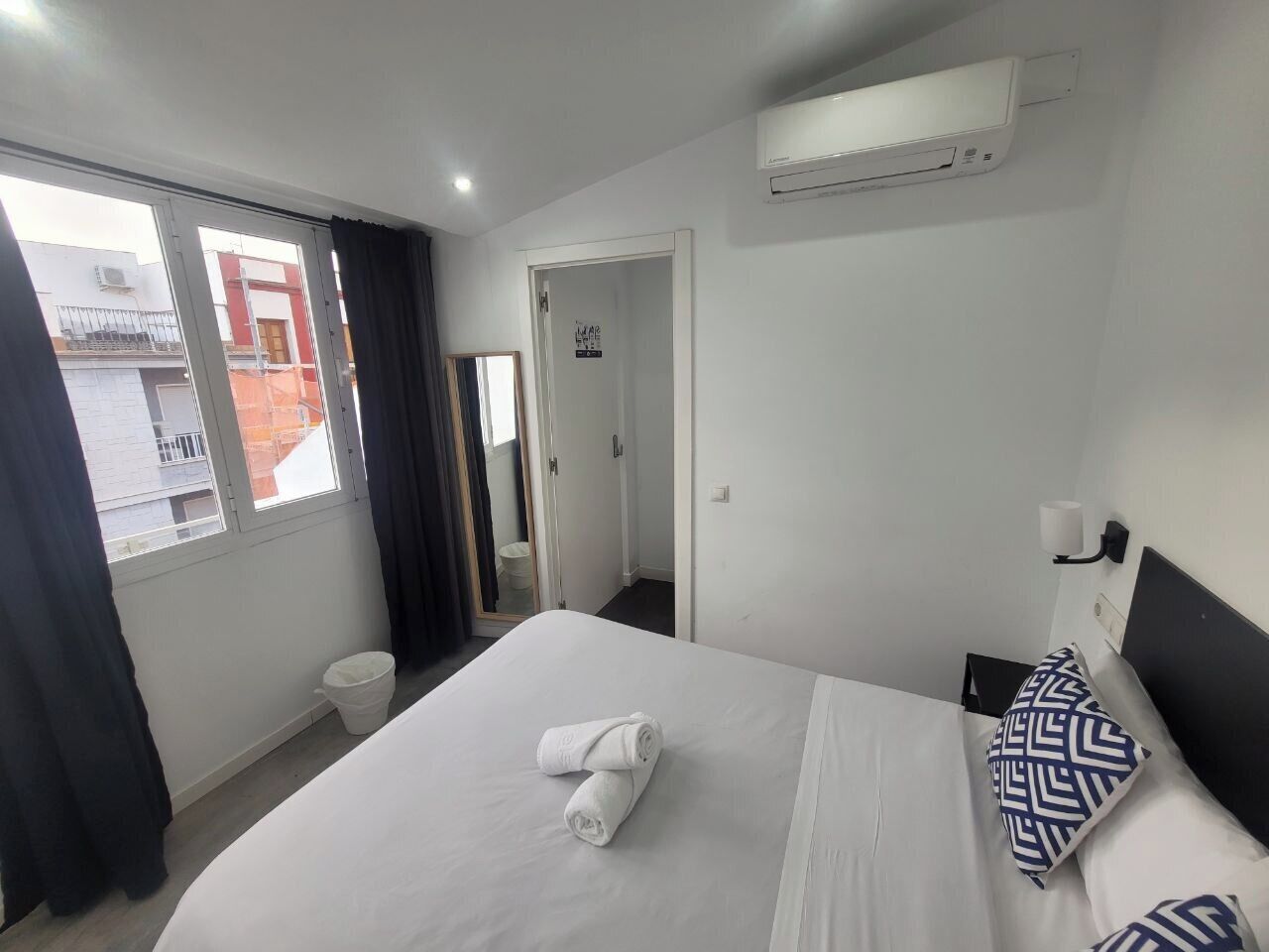 A hotel room with a large bed, a window with curtains, a white air conditioning unit, and a door leading to another room.