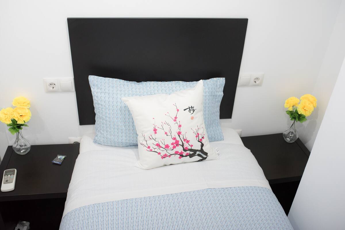 A neatly made bed with a white and blue patterned bedspread, a pillow with a tree design, and two vases with yellow flowers on either side.