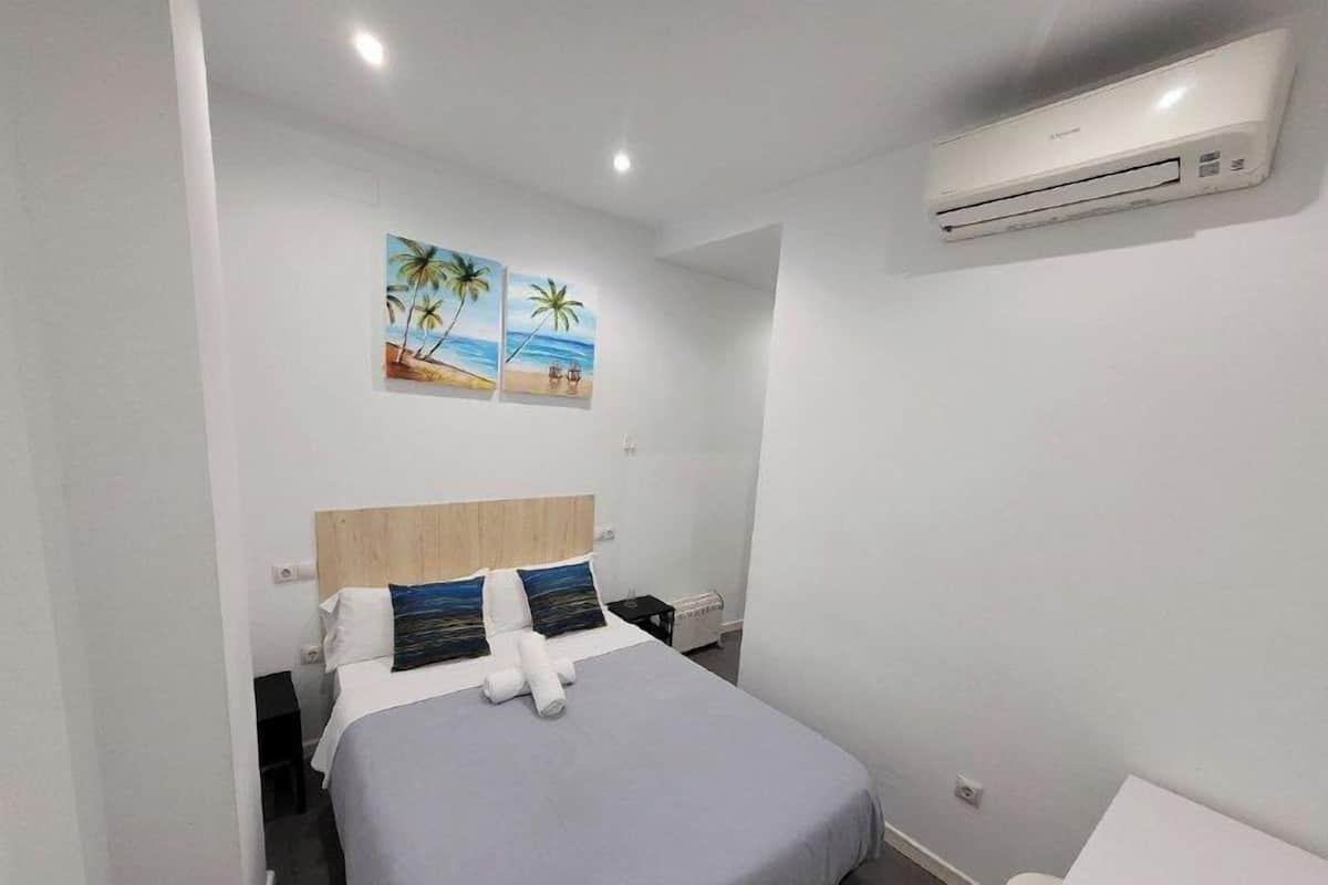A hotel room with a bed, a small table, an air conditioning unit, and two wall paintings of a beach scene.