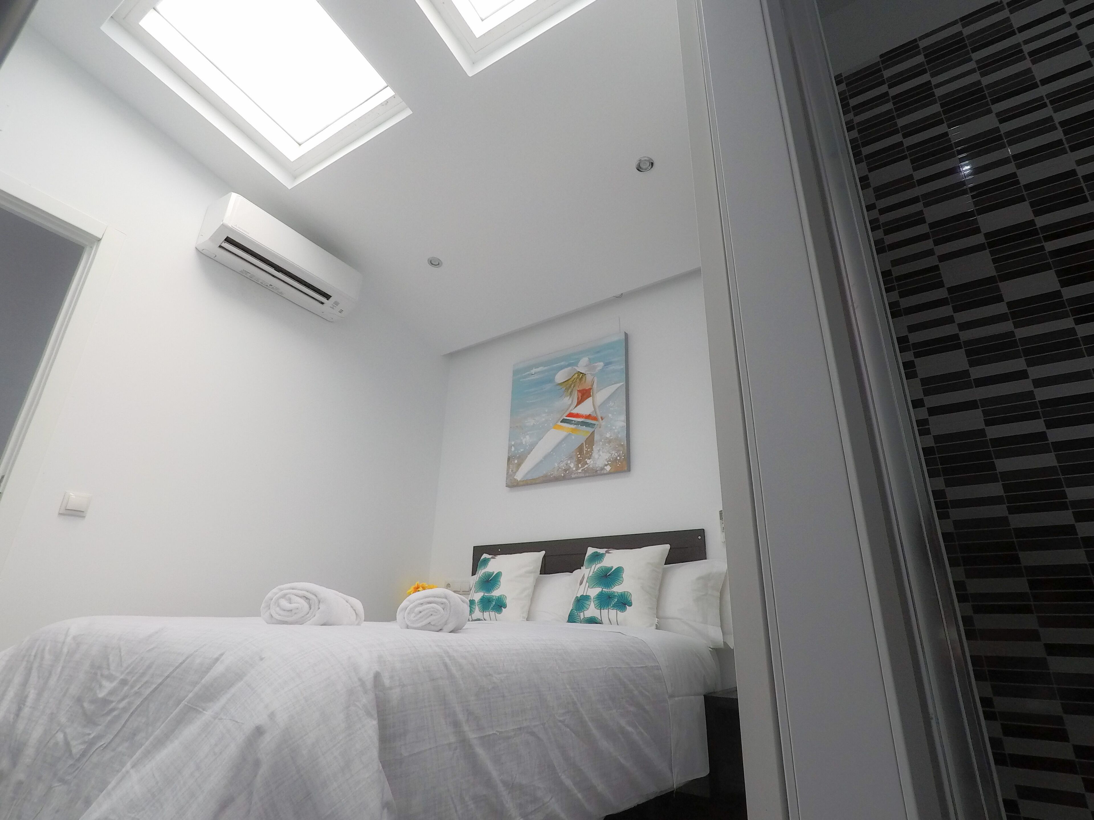 Habitación de hotel con cama, ropa de cama blanca, un cuadro de una playa y tragaluces.