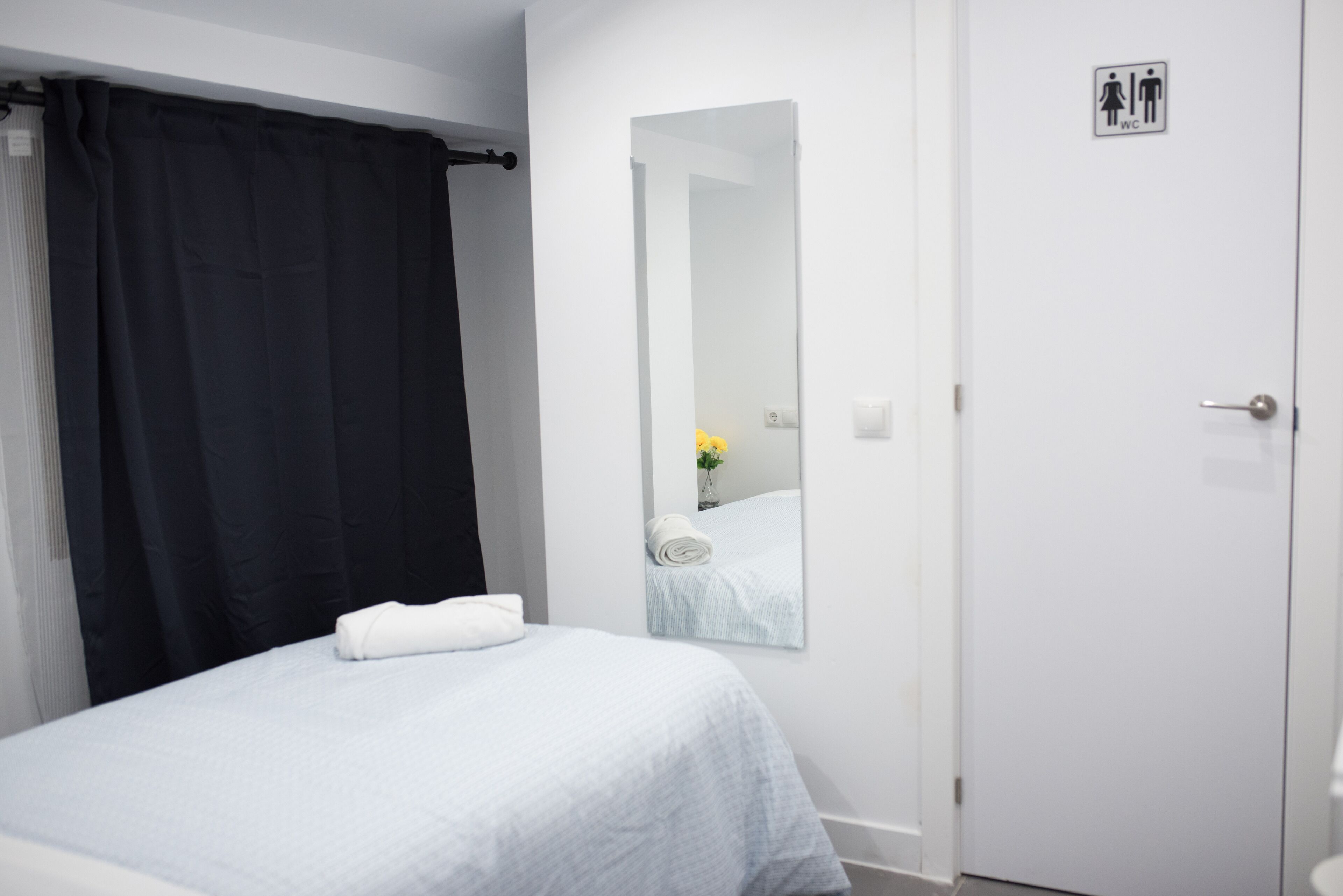 Habitación de hotel con cama, cortina negra, puerta blanca y espejo que refleja una toalla y un florero.