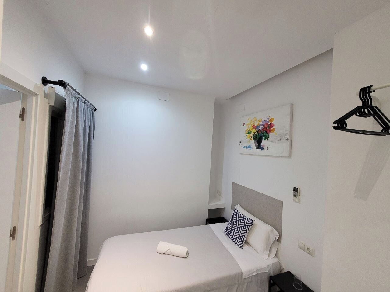 A neatly arranged hotel room with a single bed, a bedside table, a hanging coat rack, and a painting on the wall.