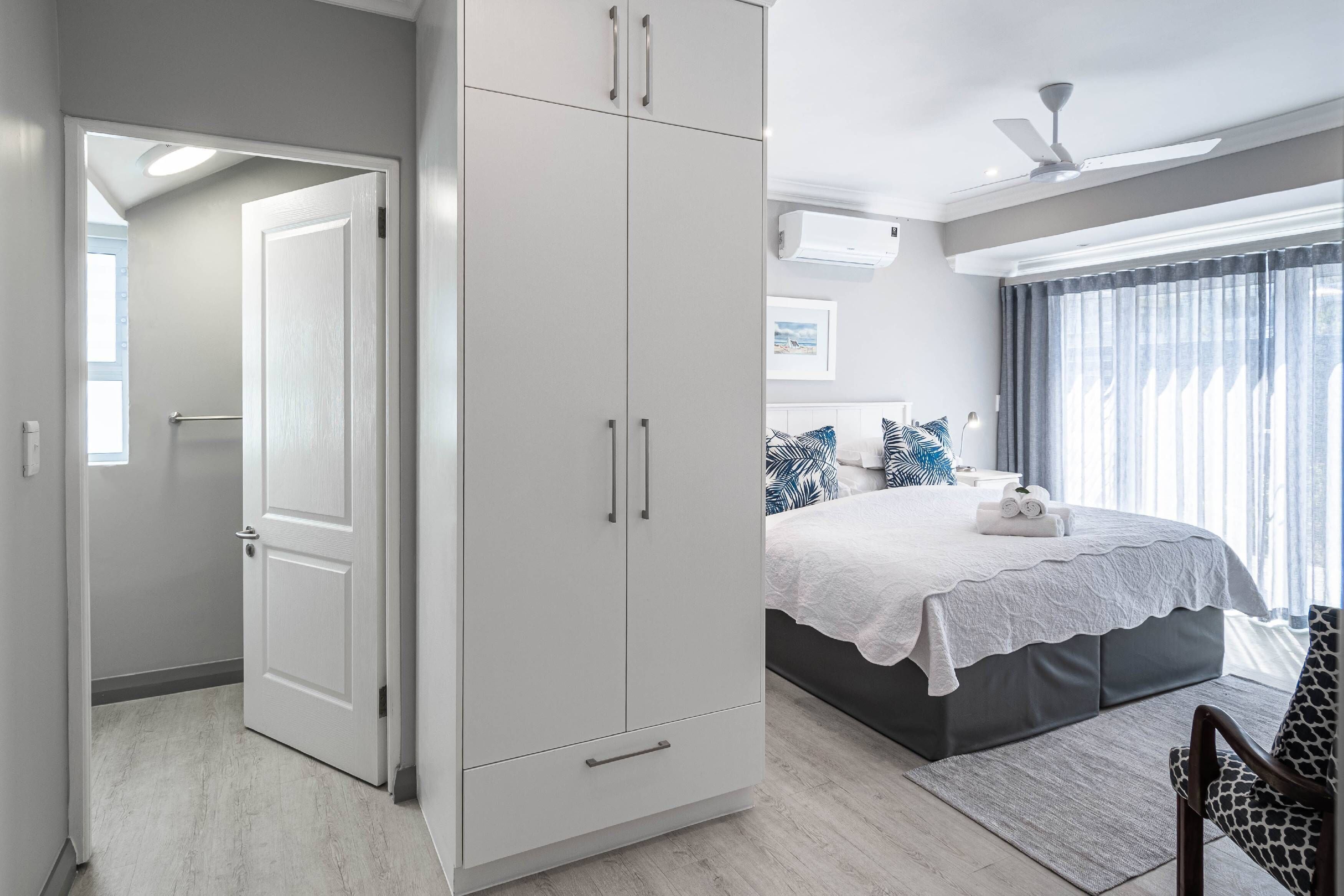 A bedroom with a large wardrobe, a bed with a white bedspread, and a view of the outdoors through a window.