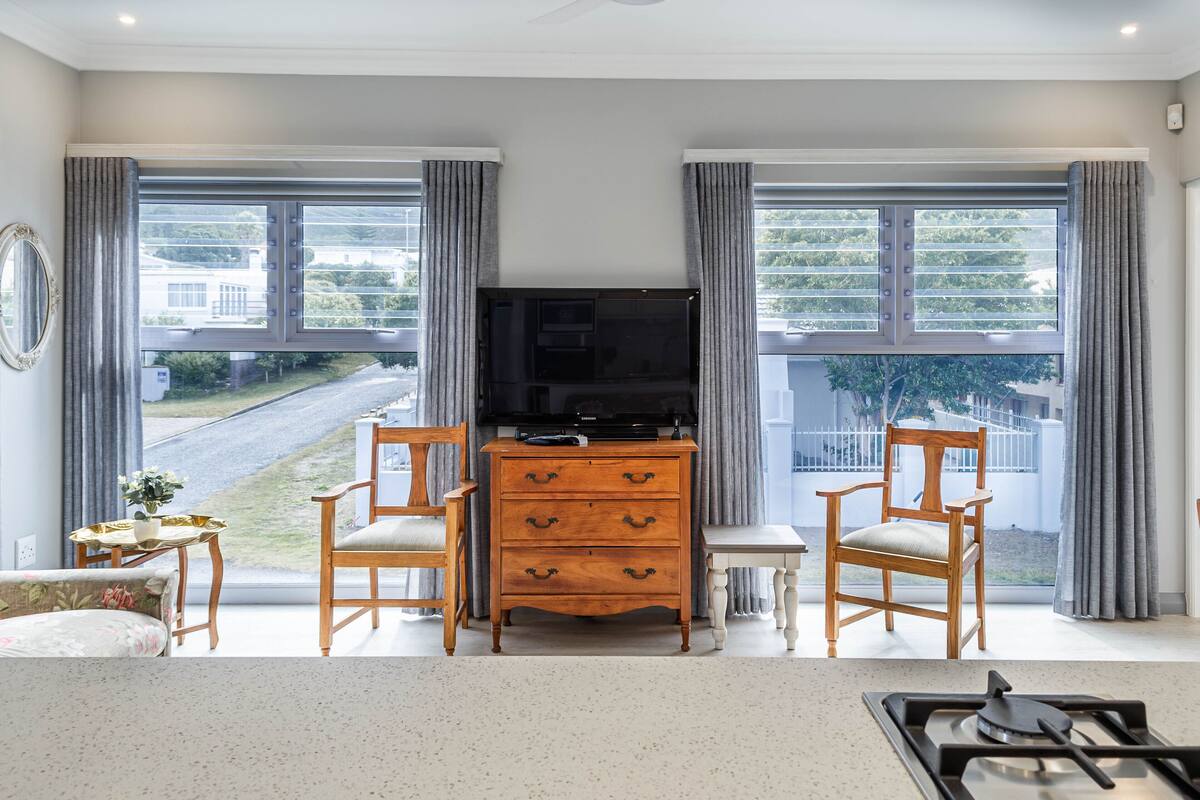 A living room with a TV, wooden furniture, and large windows with curtains.