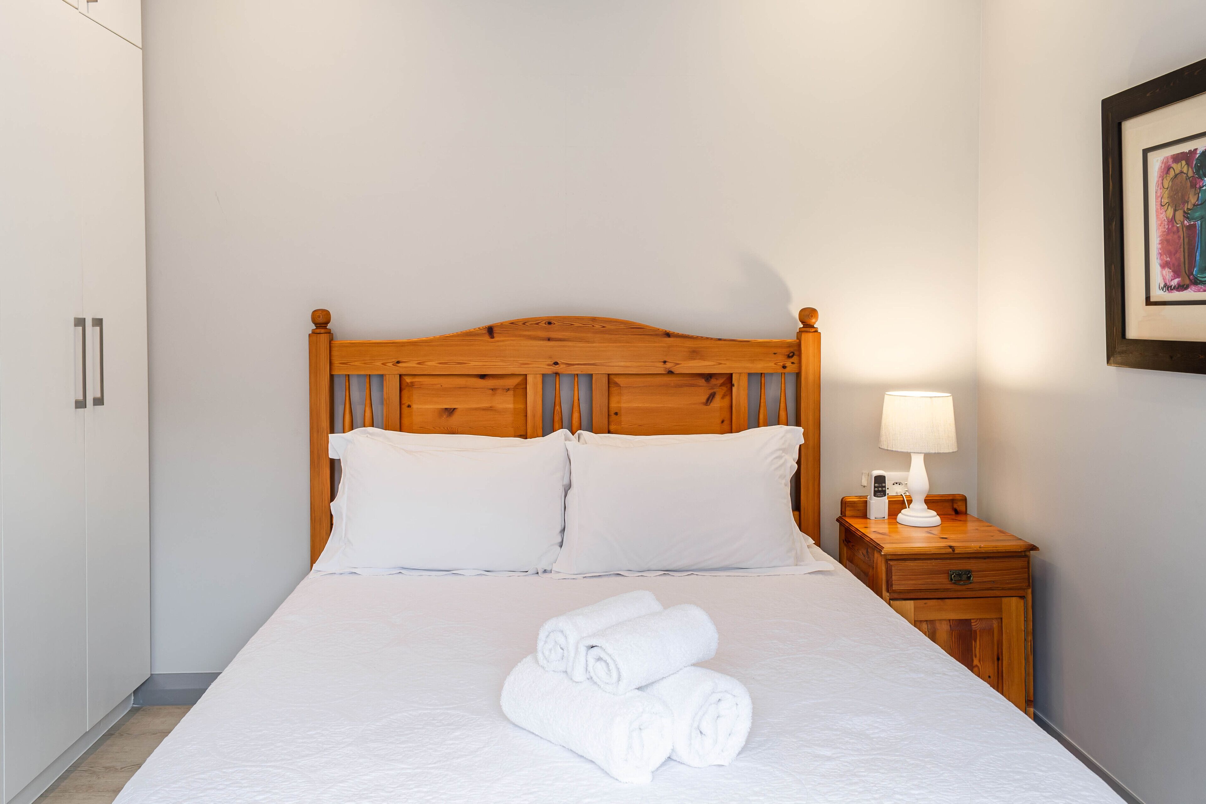 A neatly made bed with white linens and rolled towels, a wooden headboard, a bedside table with a lamp, and a framed picture on the wall.