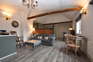 Uma sala de estar aconchegante com um sofá cinzento, cadeiras de madeira, uma pequena mesa e uma escada que leva a outro quarto.
