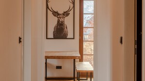 Ein Flur mit einem Hirschbild in einem Rahmen, einer Holzbank und einer Tür, die in einen Raum mit einem Fenster und Blick auf Herbstbäume führt.