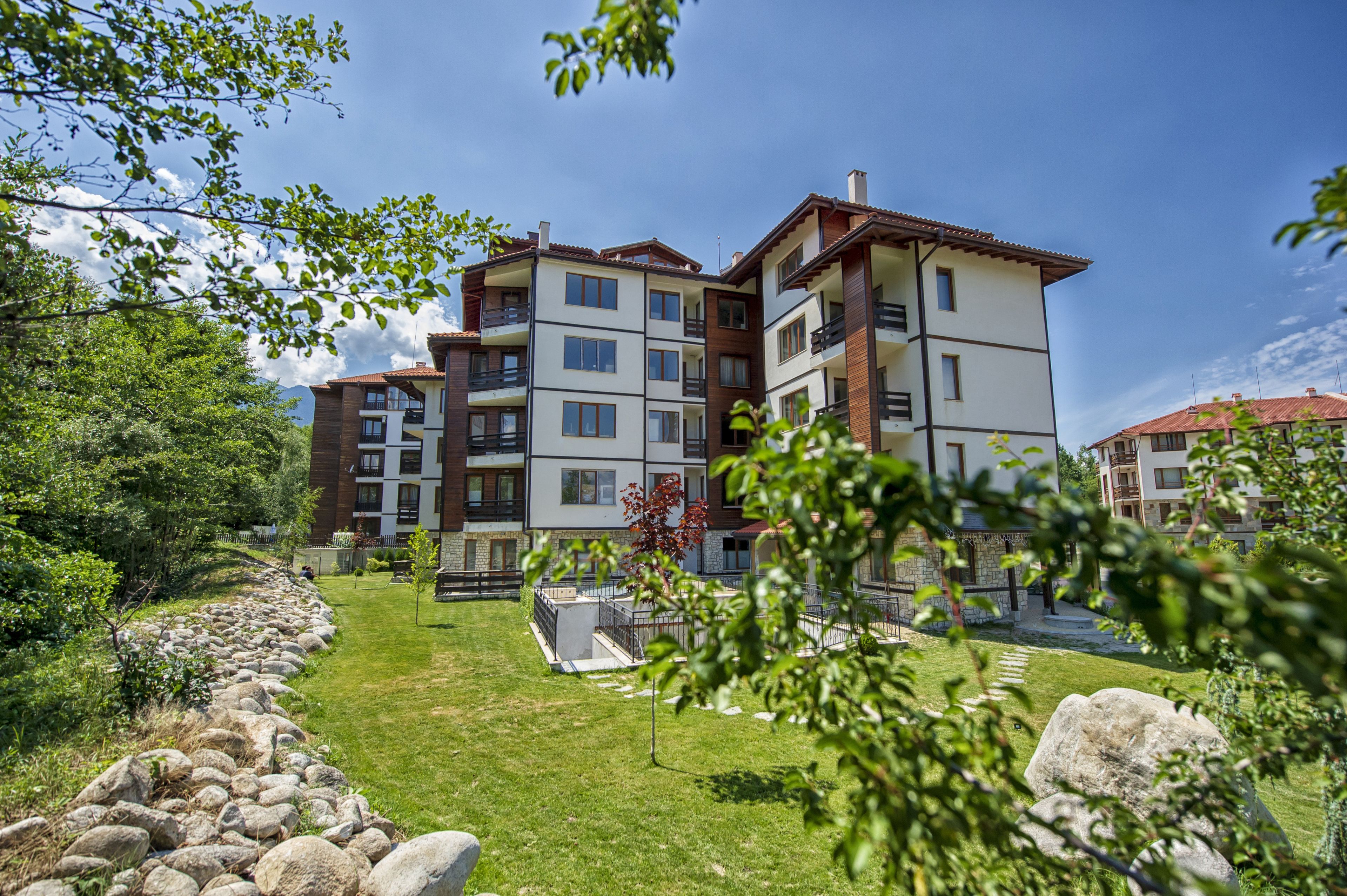 A multi-story residential building with balconies, a stone pathway, and a grassy lawn.