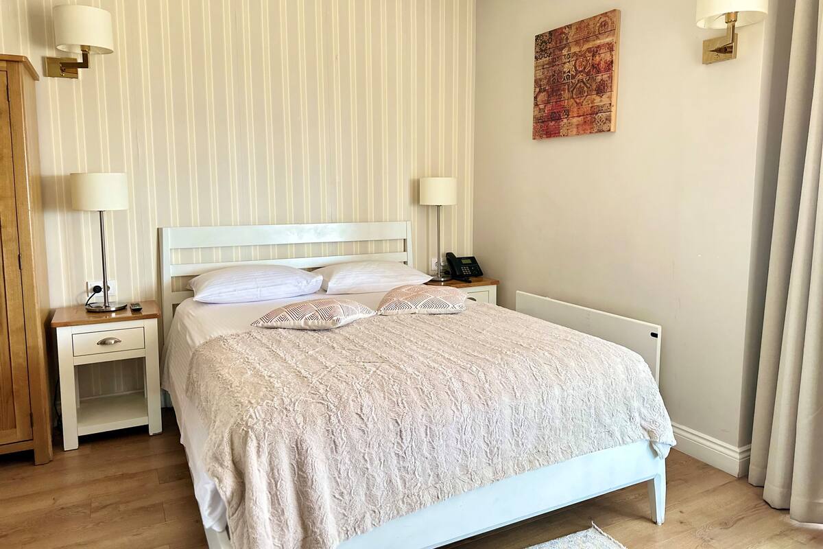 Una cama bien hecha con cabecera blanca, dos mesitas de noche con lámparas, un teléfono junto a la cama y una obra de arte decorativa en la pared.